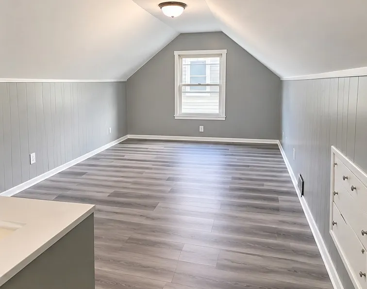 Attic bedroom after the remodel: smooth white sloped ceiling, gray painted paneling, new plank flooring and a window at the far end