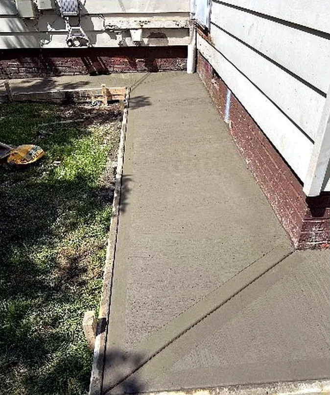 Side of the house after the work: a freshly poured concrete walkway along the foundation, still in its wooden forms