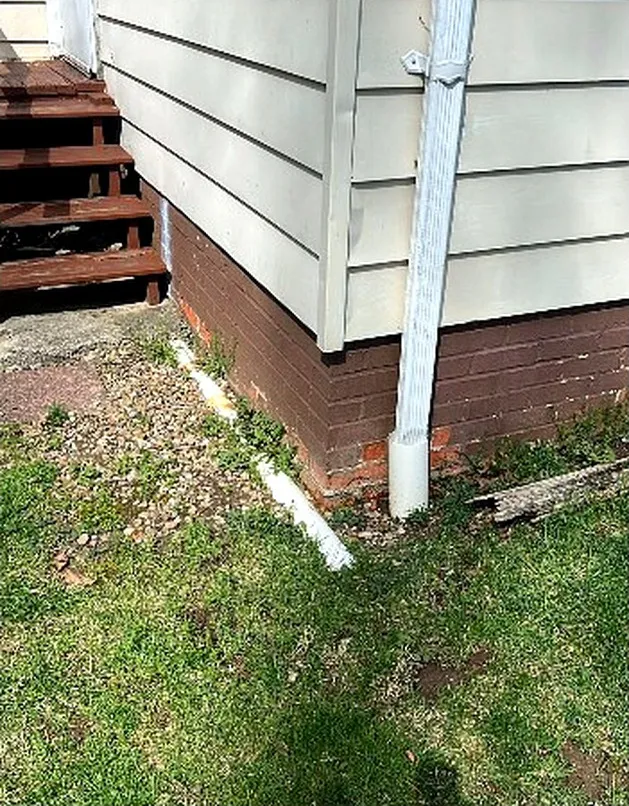Side of the house before the walkway: grass, gravel and a downspout along the brick foundation