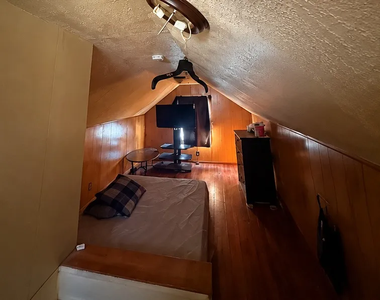 Attic bedroom before the remodel: textured sloped ceiling, dated wood paneling, worn wood floor and old furniture